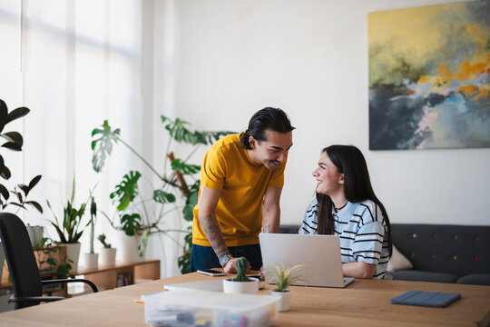 A young man and woman work together in a bright office. He stands beside her desk, leaning in to look at her laptop screen while they chat and smile.