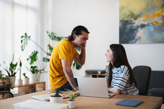 A young man wearing a yellow shirt leans over a wooden desk to converse with a woman working on a laptop. They are collaborating together in a bright, modern office space.