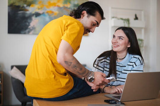 A young man in a yellow shirt and a woman with dark hair smile while looking at a smartphone together. They are sitting at a table with a laptop in a bright modern living room.