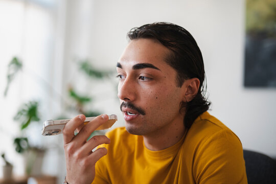 A young man with dark hair and a mustache wearing a yellow shirt holds his smartphone horizontally close to his mouth to record a voice message while sitting indoors.