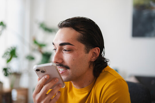 A young man with a mustache and dark hair is smiling while recording an audio message on his smartphone. He wears a yellow t-shirt and sits indoors near plants.