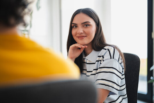 A young brunette woman sits across from a colleague in a bright office. She rests her chin on her hand and smiles warmly while listening to the conversation during a meeting.