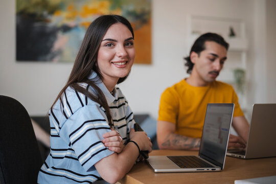 A young woman smiles warmly while sitting at a wooden desk with her laptop. In the background, a man works on his computer in a bright home office setting.