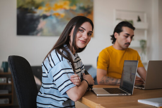 A young woman with dark hair sits at a wooden table with her laptop, smiling warmly at the camera while a male colleague works remotely in the bright home office.