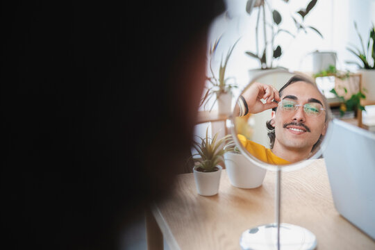 A young man with a mustache wears a yellow shirt while sitting at a desk. He looks into a round tabletop mirror, smiling and adjusting his glasses in a bright room.
