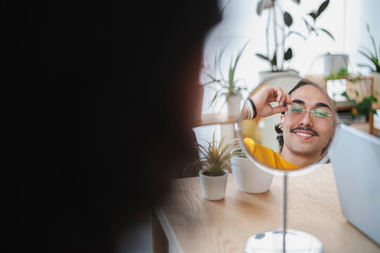 A young man with a mustache is smiling while adjusting his glasses in a round mirror on a wooden desk. He is indoors with plants in the background during the day.