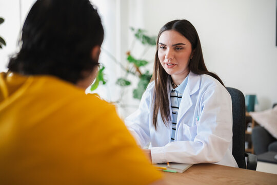 A young female doctor in a white coat is consulting with a patient in her office. She is talking and explaining a medical diagnosis during the appointment.