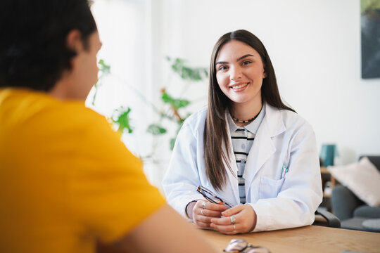 A young female doctor wearing a white lab coat sits at a wooden desk, smiling warmly at a male patient during a medical consultation in a bright clinic office room.