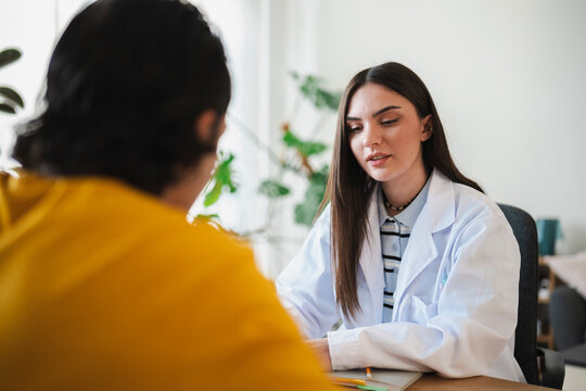 A young female doctor in a white lab coat sits at a desk and consults with a patient in an office. They discuss medical treatment during a private healthcare appointment.