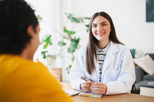 A smiling female doctor in a white coat sits at a desk during a consultation with a patient in an office. They are discussing health treatment options in a modern clinic.