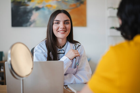 A smiling female doctor wearing a white coat sits at her desk during a medical consultation with a patient in an office. A mirror and laptop rest nearby on the table.