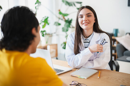 A smiling female doctor in a white coat sits at her desk in an office, holding glasses while consulting with a male patient sitting across from her during an appointment.