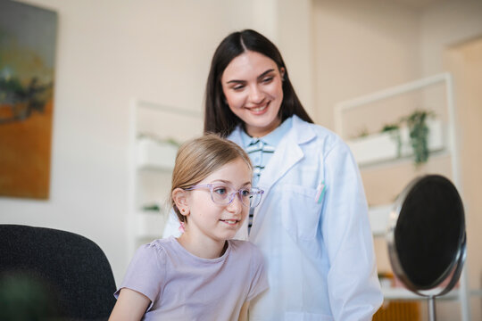 A young girl wearing new eyeglasses looks into a mirror during an eye exam. A smiling female optometrist stands behind her in the clinic, guiding the vision test.