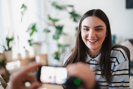 A young woman with long dark hair smiles cheerfully while an unseen person records a video of her using a smartphone mounted on a small tripod in a bright room.