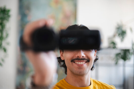 A young man with a mustache and a yellow t-shirt is smiling while holding a black video camera up to record a home video inside a bright modern living room.