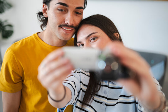 A happy young couple stands close together, smiling cheerfully while recording a video of themselves with a small vintage camcorder held by the woman in their living room.