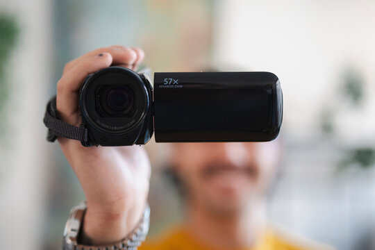 A person holds a black digital video camera up toward the camera lens, recording a video indoors. The device is held steady in a hand while the background remains blurred.
