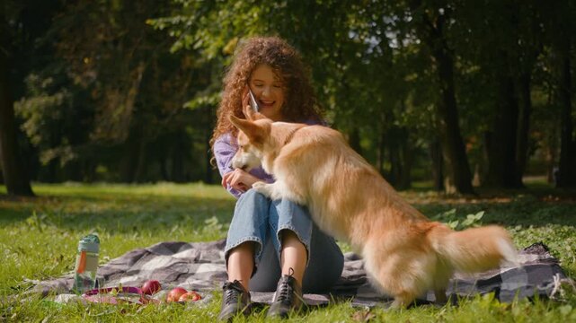 Smiling Caucasian gen z girl chatting on smartphone in park with fluffy corgi dog playing outside happy pretty 20s young woman talking mobile phone call female with domestic pet puppy friend in nature