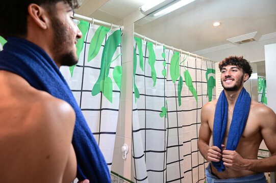 Handsome young man, feeling confident looking himself at bathroom mirror