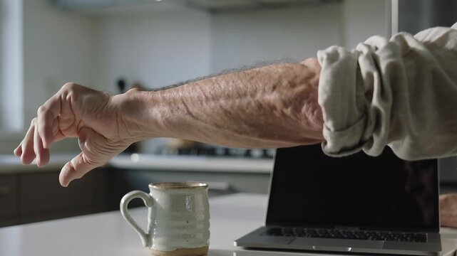 Resting senior arm rolled sleeve testing rotating wrist, curling fingers in kitchen, mug, laptop