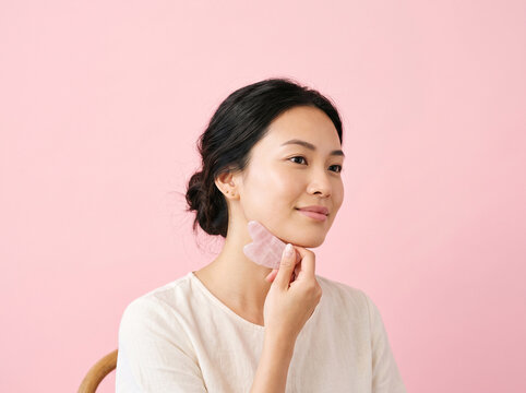 Asian woman using a rose quartz gua sha facial massage tool on her jawline, beauty and skincare concept, copy space