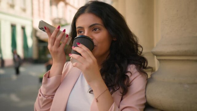 Happy young Indian business woman listening voice message on phone outside city Arabian businesswoman girl female worker drink cup coffee manager receiving audio use cellphone social media town street
