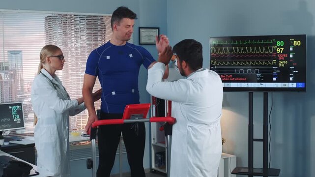 Two medical professionals apply electrodes on a muscular male athlete standing on testing equipment inside a modern medical laboratory with urban skyscrapers visible outside.