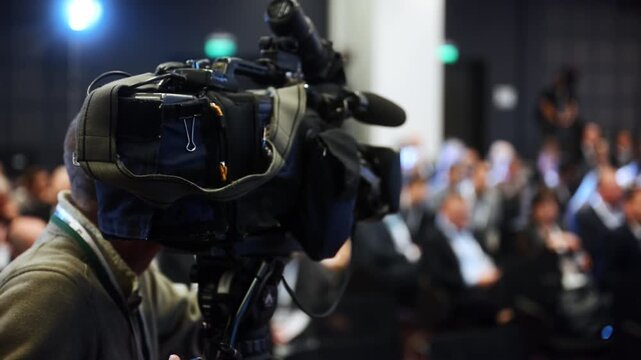 Professional video camera filming a large business conference with attendees in the audience. Concept of event coverage, media, communication and corporate presentation.