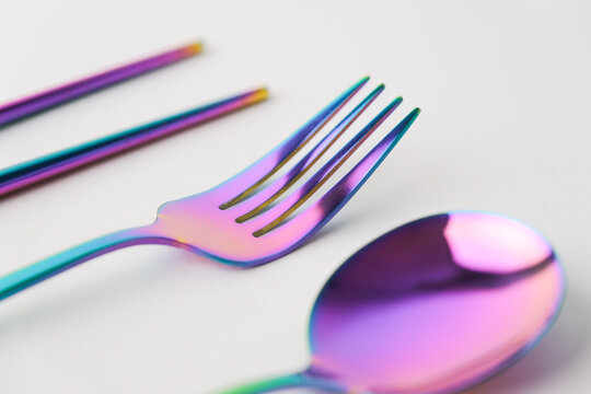 iridescent metallic sticks , fork and spoon resting on a glossy white surface, vibrant rainbow gradient reflecting soft pink and blue light
