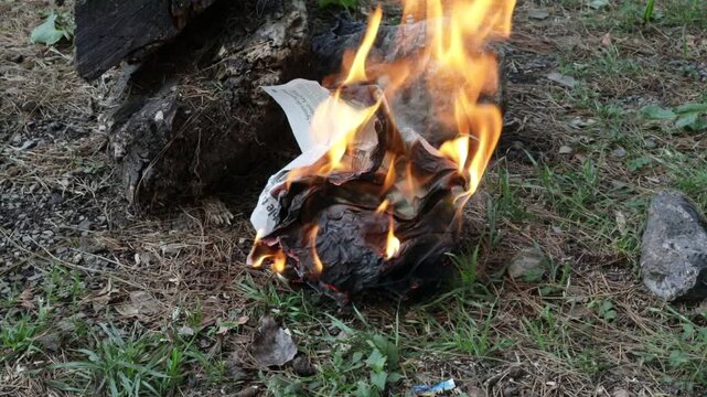 A close-up shot of a crumpled piece of printed newspaper catching fire and burning on the ground outdoors.