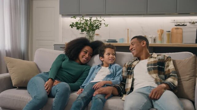 Relaxed tired African American family dad mom son falling on sofa living room at home rest celebrating couch comfortable relocation moving rented adult parent female male little child kid indoor flat
