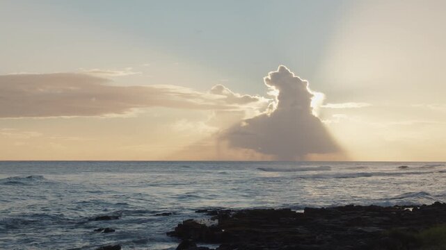 Sunbeams stream behind a cloud above waves and a rocky coast in Kauai, Hawaii.
