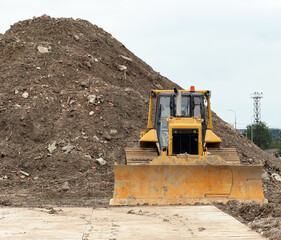 Heavy industrial yellow crawler bulldozer working at a construction site. Powerful earthmoving machinery vehicle clearing ground near a huge pile of soil dirt and stones under an overcast sky.  © Dmitry Presnyakov