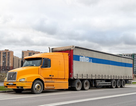 Classic orange Volvo VNL bonnet semi-truck tractor unit with a large cargo trailer parked on a city street road. Big American style Swedish commercial vehicle under a cloudy sky. Residential block.