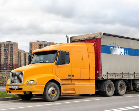 Classic orange Volvo VNL bonnet semi-truck tractor unit with a large cargo trailer parked on a city street road. Big American style Swedish commercial vehicle under a cloudy sky. Residential block.