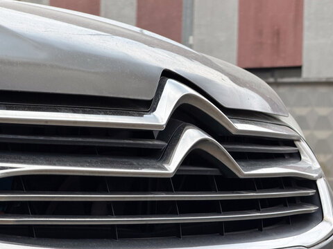 Chrome Citroen double chevron logo close-up on a grey metallic car radiator front grille. French automobile corporate brand emblem suitable for vehicle models like C4, C5, C3, C5 X, Berlingo, Jumpy.