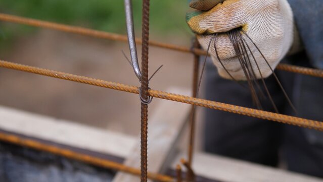 Close-up of gloved hand fastening rusty reinforcing steel bars with tie wire on building site, showing manual rebar binding during concrete reinforcement work with shallow focus and industrial texture