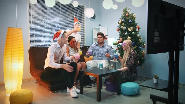 Five young adults wearing Santa hats toast drinks in a modern indoor lounge decorated for Christmas during daytime. They celebrate together near a decorated tree.