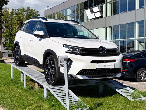 White Citroen C5 Aircross compact SUV crossover car displayed on a metal ramp platform outdoors next to a modern dealership showroom building facade with glass windows under a blue sky with clouds.