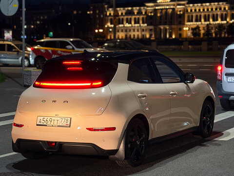 Stylish modern Ora 03 Good Cat electric hatchback car parked on a city street at night. Premium Chinese EV automobile with a glowing red LED taillight strip, neo-retro design, and two-tone body color.