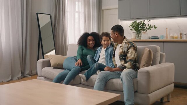 Excited happy African American family mom dad son bringing new sofa to apartment smiling ethnic woman little boy strong man moving couch furniture mother father sitting hugging enjoying rest home