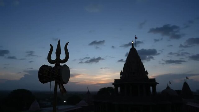 A golden trident and small drum stand against a historic temple at dusk