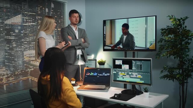 A team of video editors collaborates in a modern office while the chief discusses a project displayed on multiple monitors during daytime.