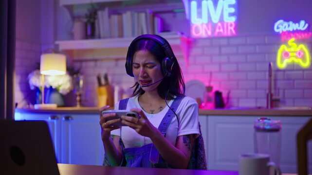 Asian young e-sport woman gamer play online mobile game on smartphone.Attractive girl vlogger influencer content creator gamer enjoy and fun live streaming broadcast games in POV screen and neon room.