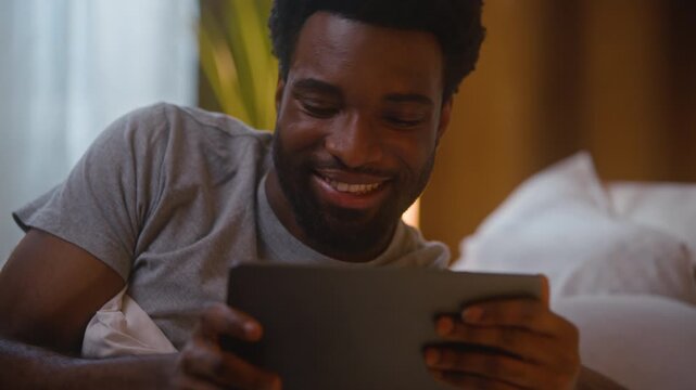 Happy smiling African American man holding digital tablet using device watching online video cheerful ethnic male guy client user browsing internet shopping lying on bed at home bedroom late evening