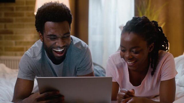 Laughing African American couple family watching comedy movie on laptop bed at home night happy man woman enjoying funny series online computer digital device relationship relaxation indoor bedroom