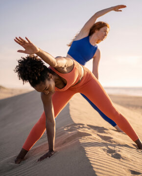 Two women of different backgrounds stretch side by side on a beach dune