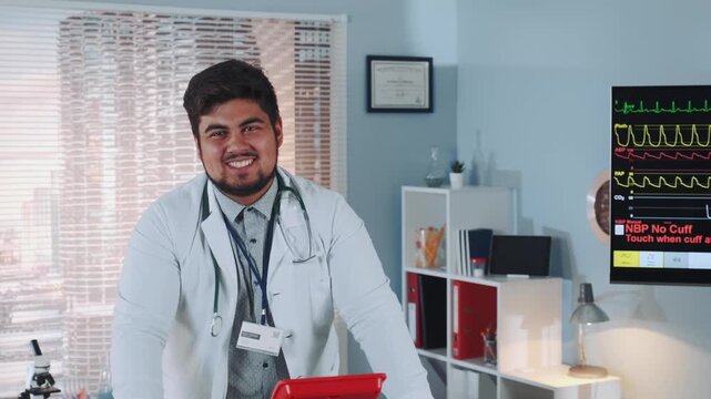 A smiling male doctor wearing a white lab coat and stethoscope holds a red tray in a medical clinic with monitors and medical supplies visible.