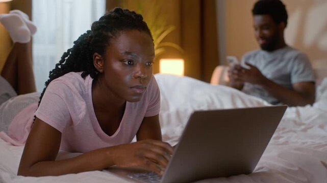 Happy family African American woman browsing internet typing laptop in bed boyfriend chatting mobile phone relaxed ethnic couple weekend online computer work gadget addiction inside home bedroom night