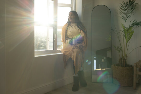 Adult woman sitting on windowsill holding ceramic mug, wearing fringed shawl and knee-high boots